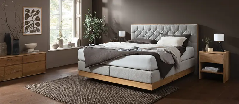 Helles Schlafzimmer mit komfortablem Boxspringbett und gepolstertem Kopfteil, Holzrahmen und passendem Nachttisch, ergänzt durch natürliche Dekoration und harmonische Farbgestaltung für gemütliche Schlafatmosphäre.